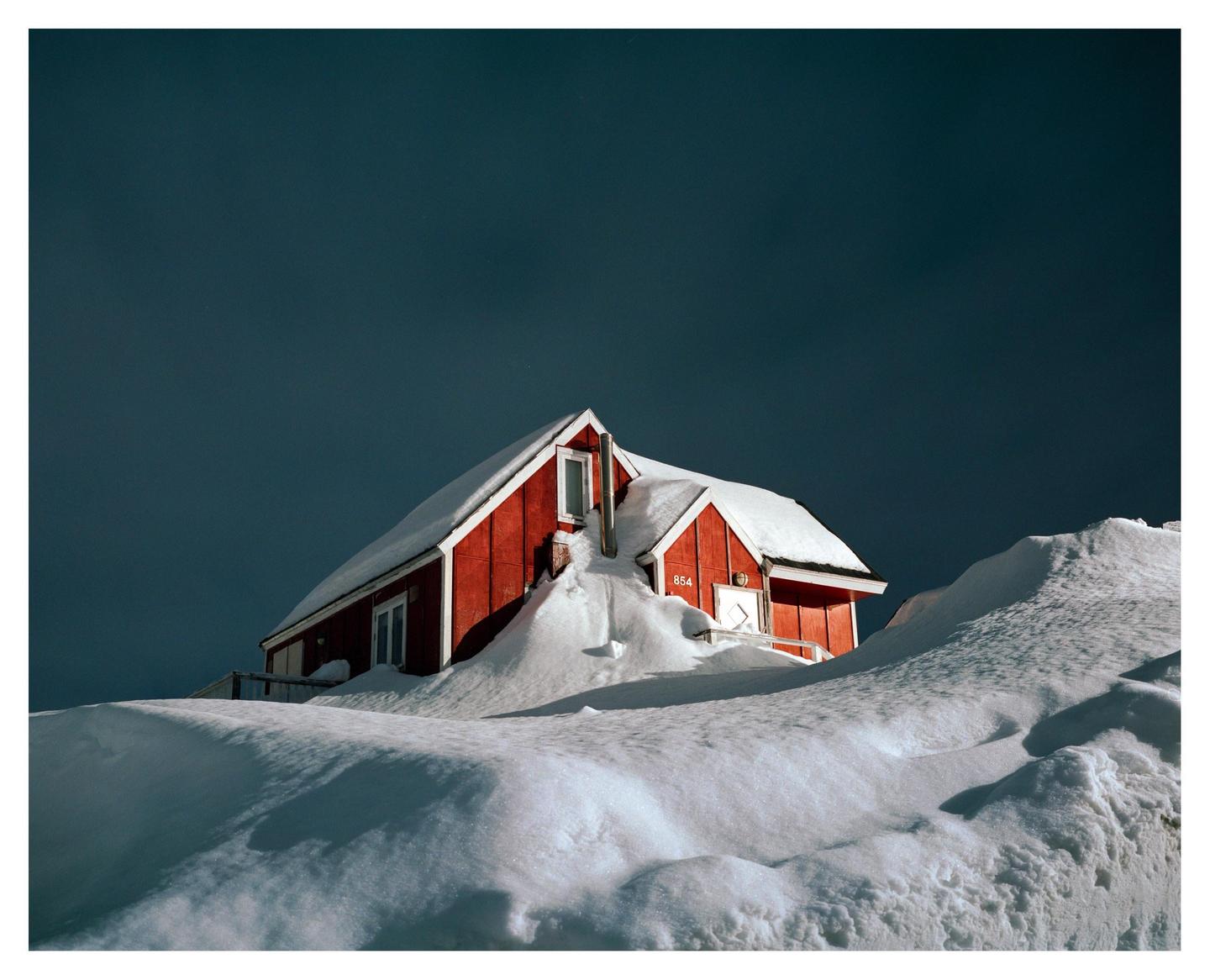 A red house with a snow-covered roof is nestled in deep snowdrifts under a dark sky.