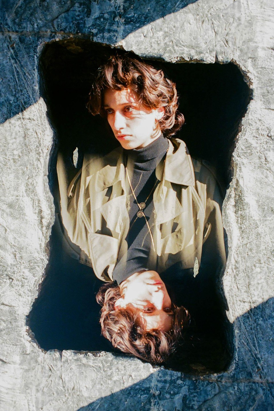 A person with curly hair and a jacket is reflected in a dark pool of water surrounded by rock.