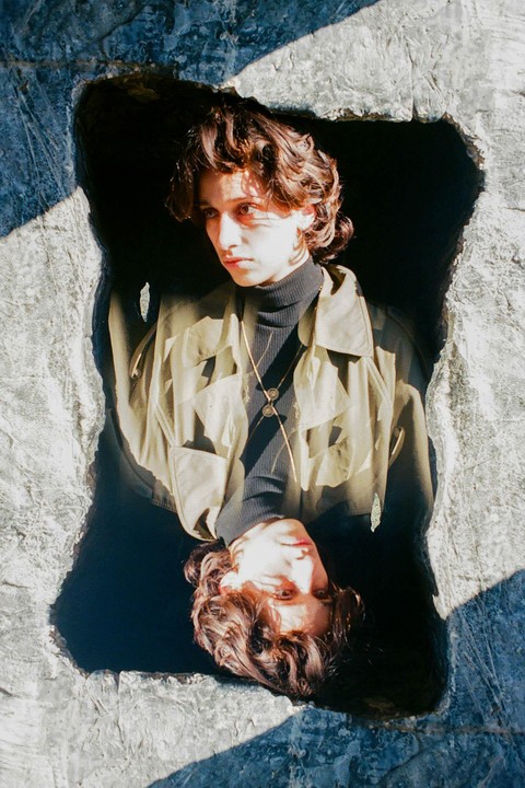 A person with curly hair and a jacket is reflected in a dark pool of water surrounded by rock.