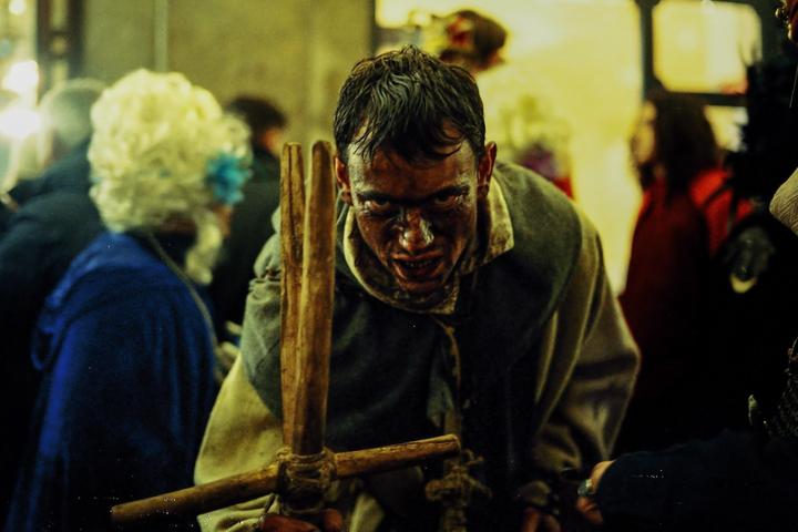 A man with a dirty face and an intense expression holds a wooden cross-like object while surrounded by people.