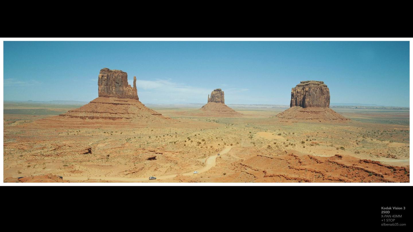 Three large rock formations stand in a vast desert landscape under a clear sky, with a dirt road winding through the foreground.