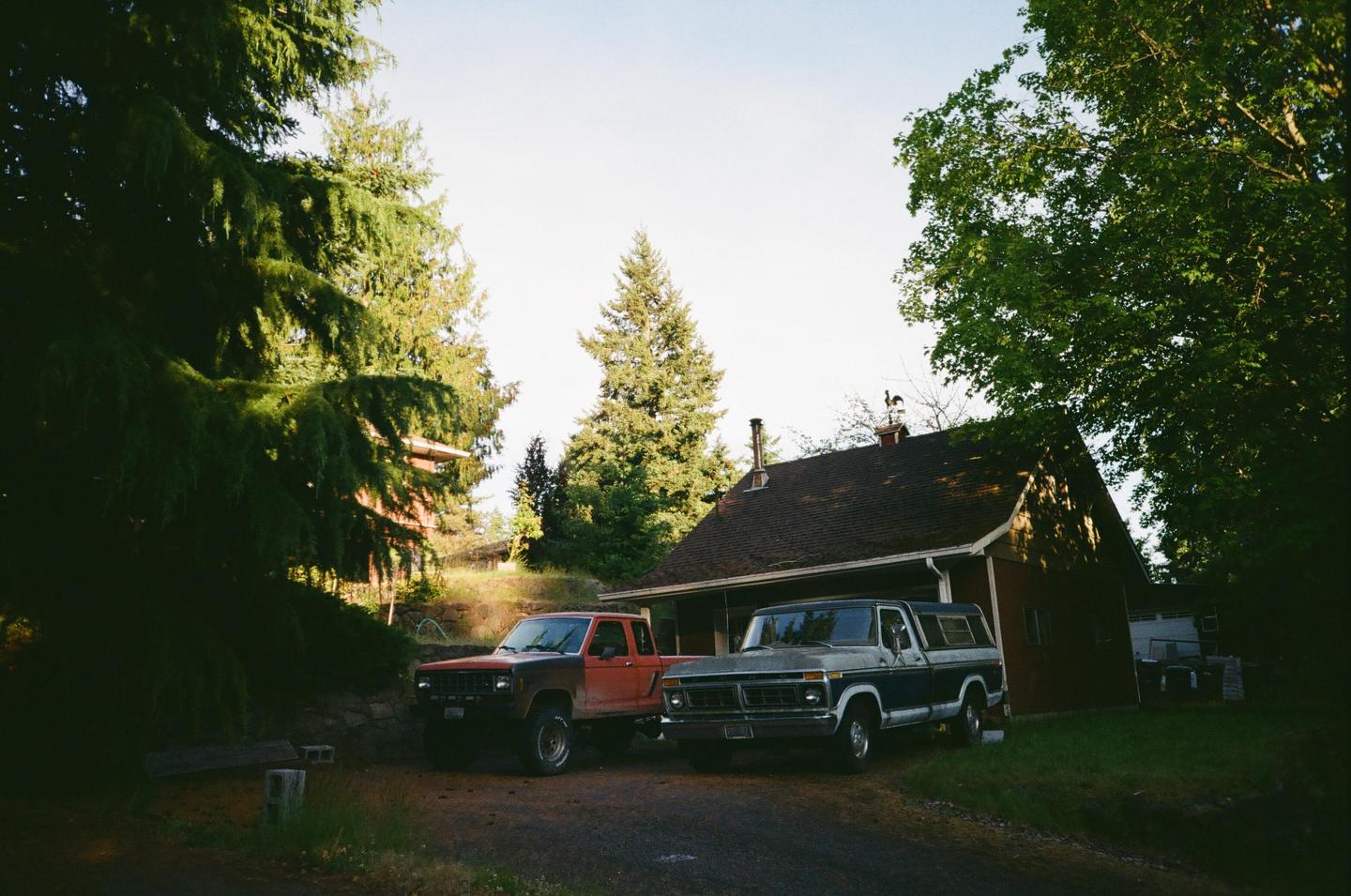 Two pickup trucks are parked on a dirt driveway next to a house surrounded by tall trees.