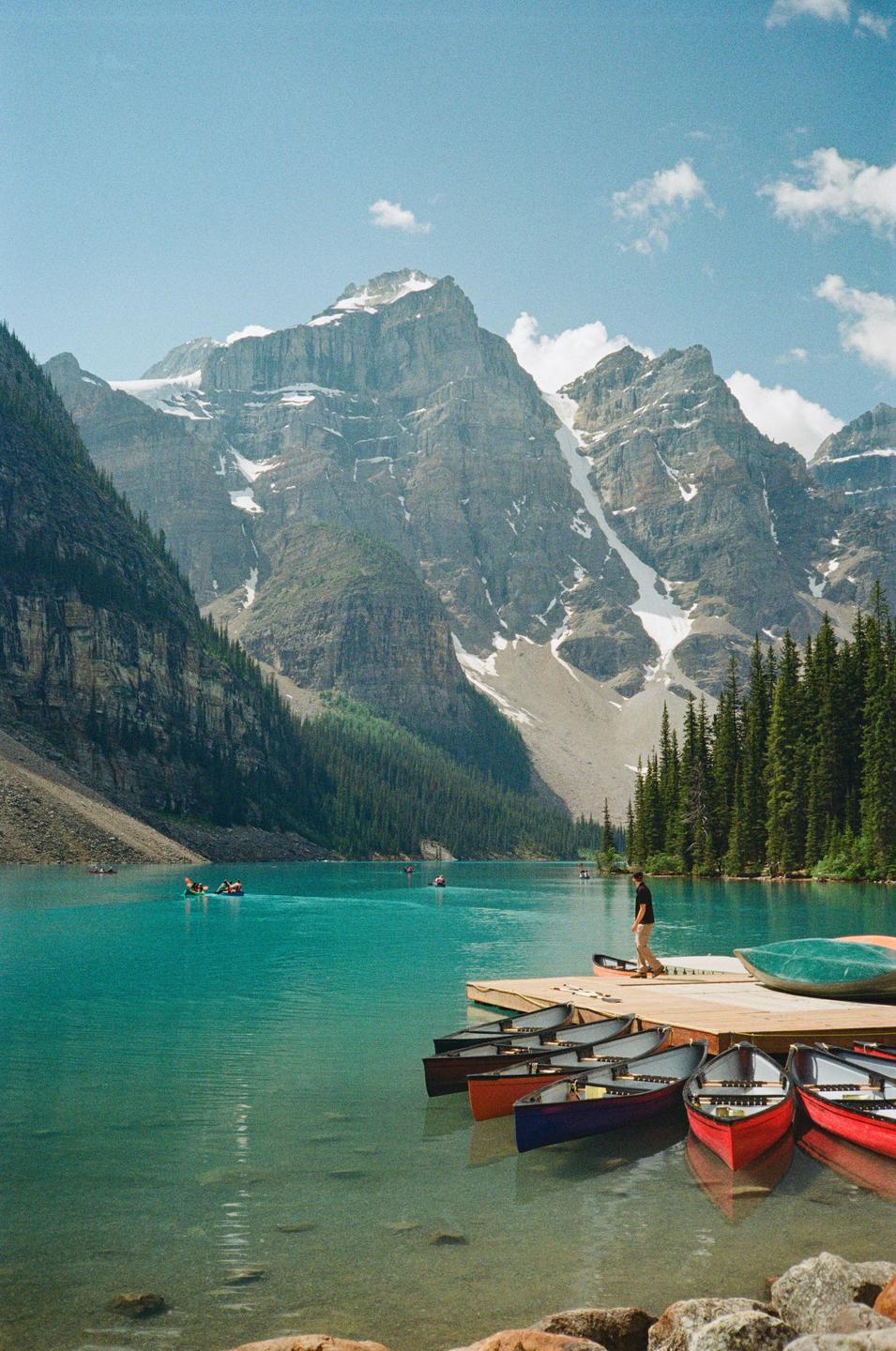 A man stands on a dock next to colorful canoes on a turquoise lake surrounded by mountains and pine trees.