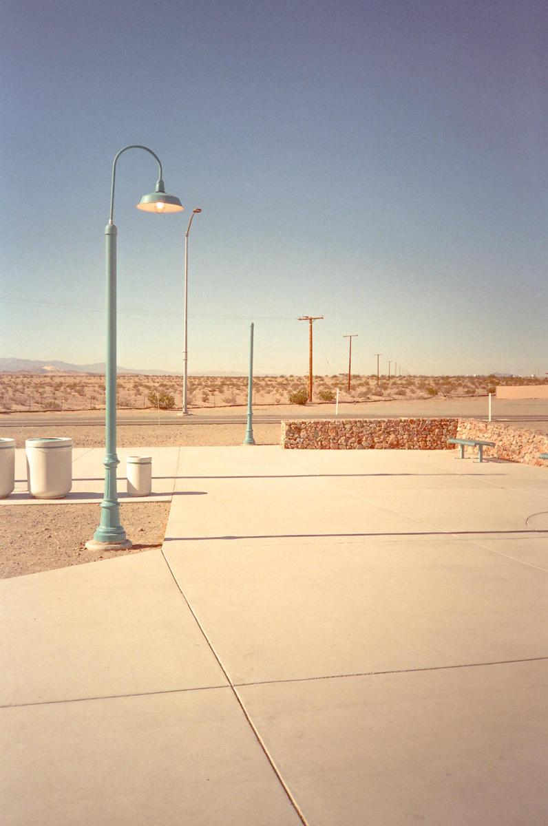 A light blue street lamp stands in the foreground of a desert landscape with a road, power lines, and mountains in the distance.