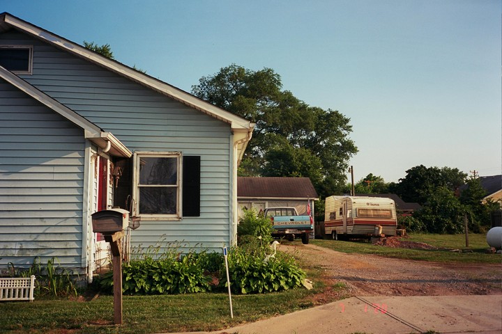 A light blue house with a mailbox in front of it stands next to a gravel driveway with a pickup truck and a camper.