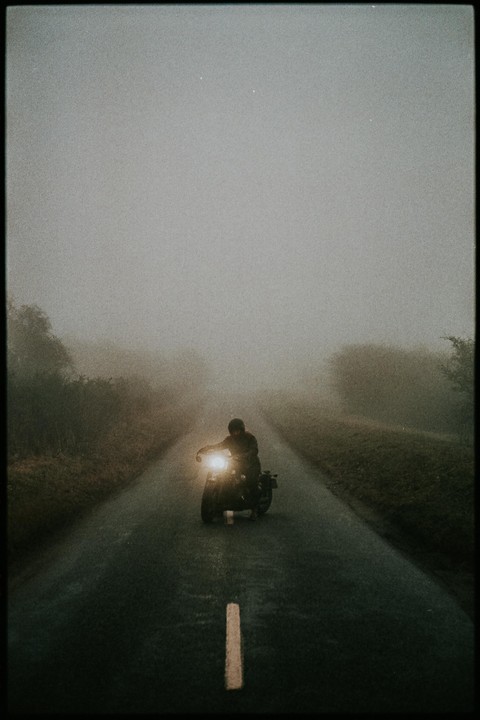 A person on a motorcycle with its headlight on drives down a road in heavy fog.