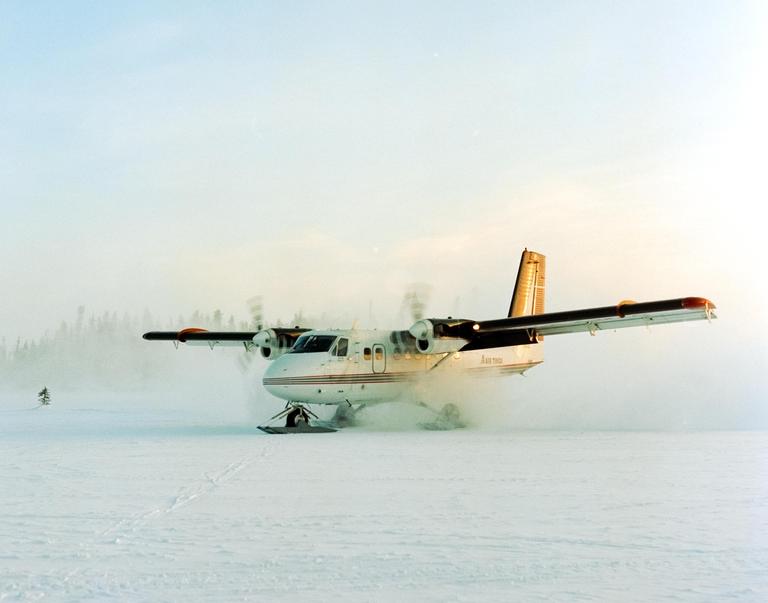 A small airplane with skis instead of wheels is on a snow-covered field with a forest in the background.