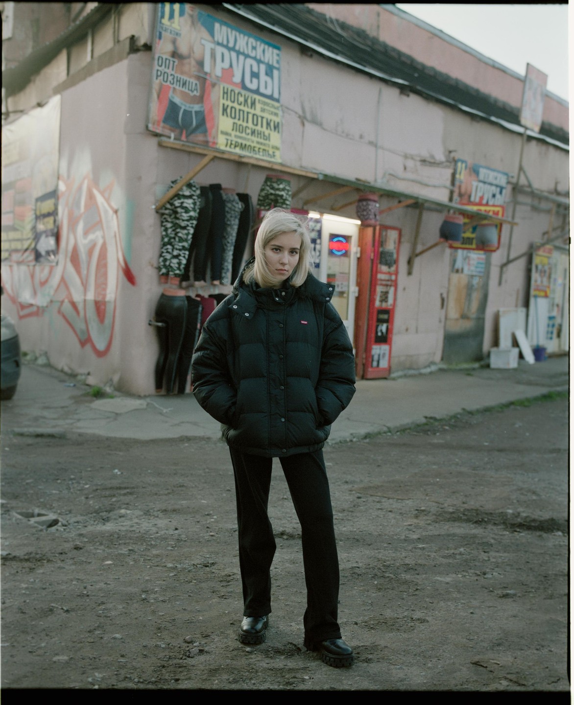 A woman stands in front of a building with signs and clothing hanging outside.