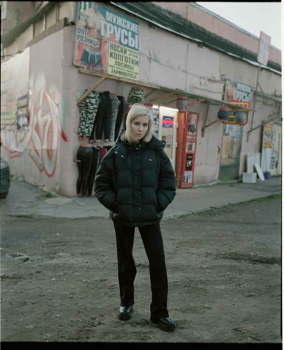 A woman stands in front of a building with signs and clothing hanging outside.
