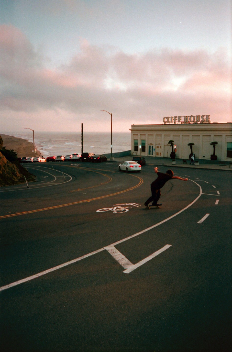 A man on a skateboard coasts down a winding road with a building and the ocean in the background at dusk.