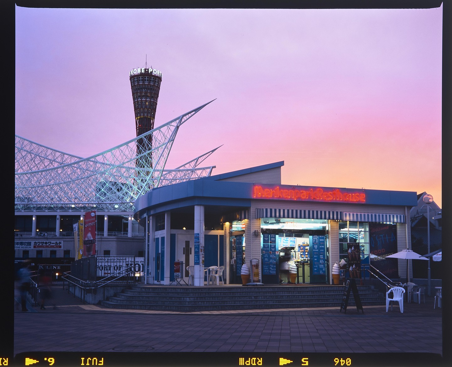 A building with a neon sign for "Merikenpark Resthouse" stands in front of a tall tower and a large white structure with a pink and orange sky in the background.