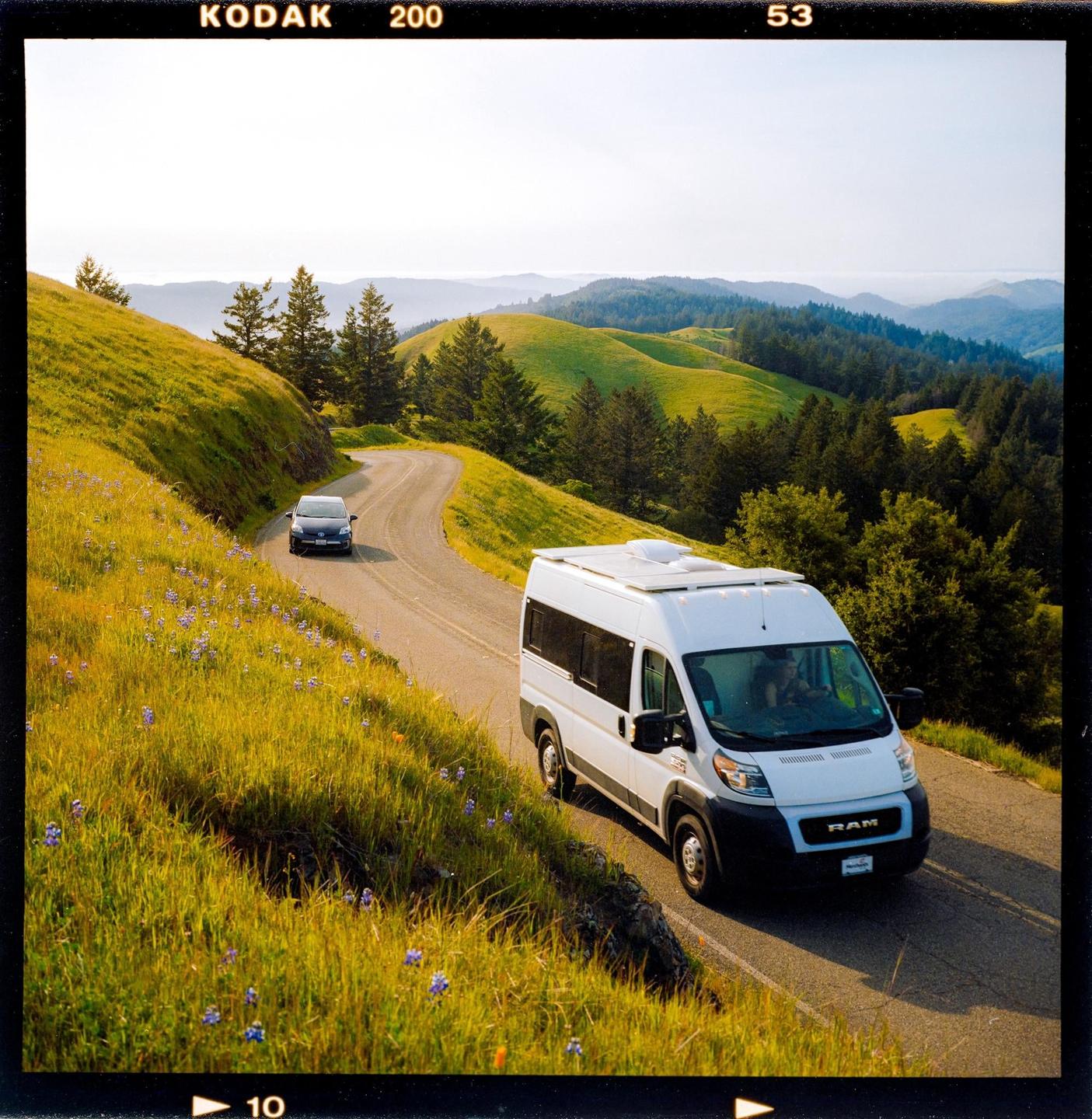 A white van and a dark car drive on a winding road through rolling green hills.