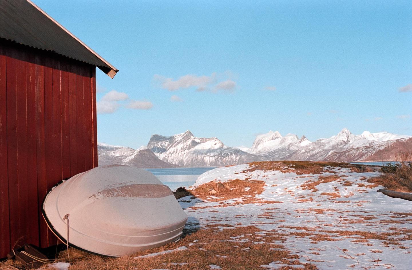 A red wooden building and an overturned boat sit on a snowy, grassy hill overlooking a body of water and snow-capped mountains under a blue sky.