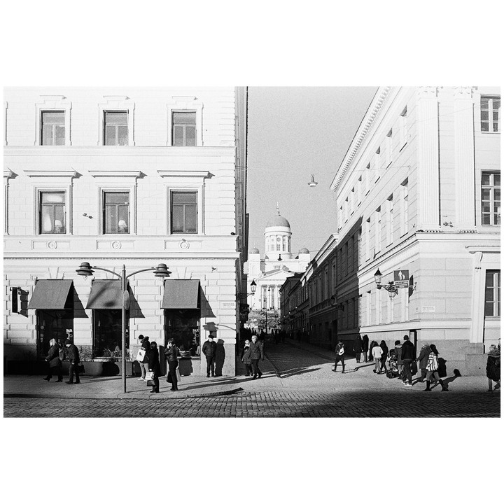 A street in Helsinki is filled with people walking on a sunny day, with a domed building visible in the distance.