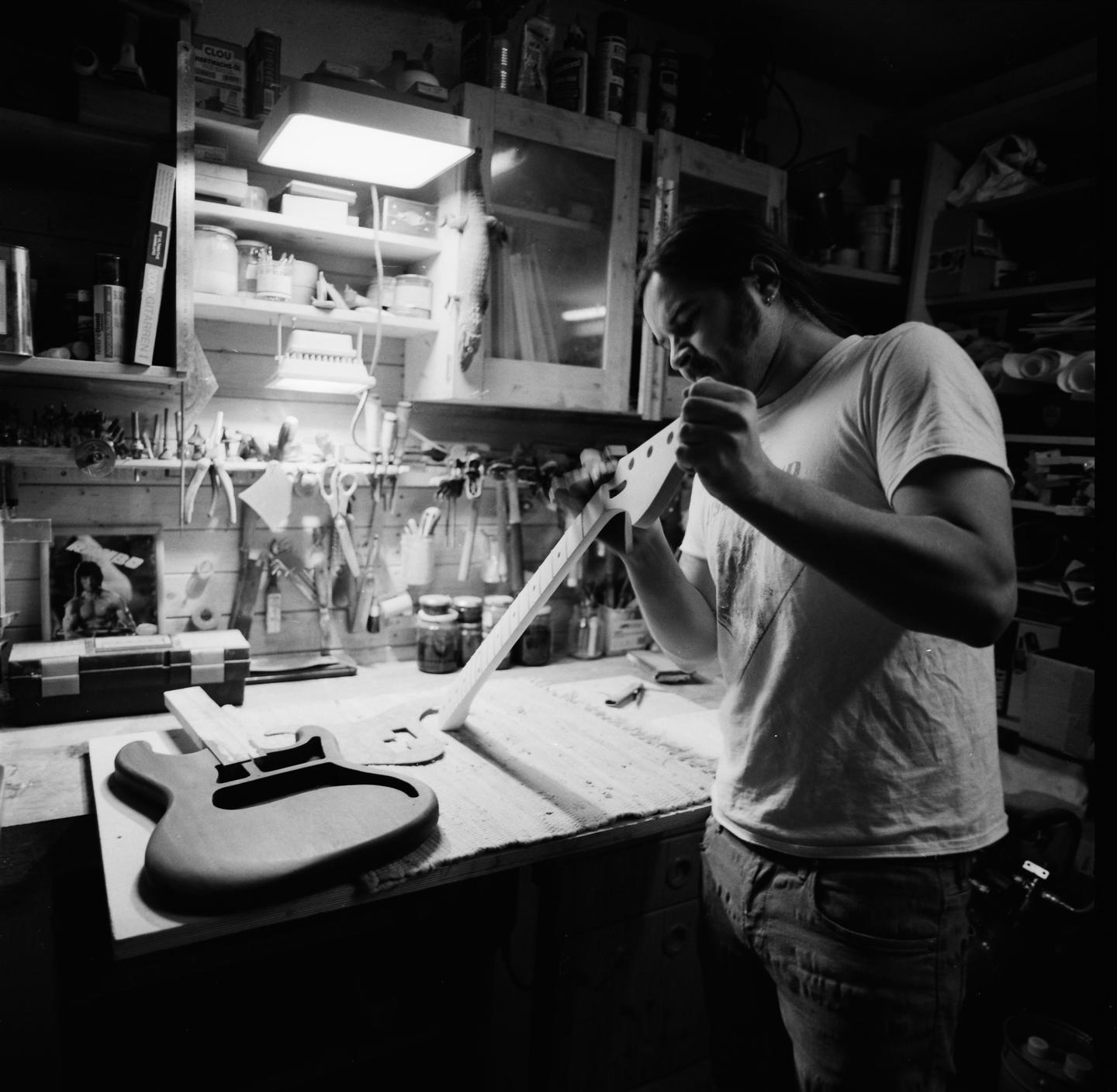 A man works on a guitar neck in a cluttered workshop.