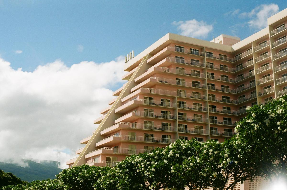 A large pink hotel with many balconies is partially obscured by flowering trees in the foreground, with mountains and clouds in the background.