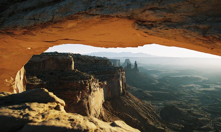A natural arch frames a vast desert landscape with rock formations and distant mountains under a bright sky.