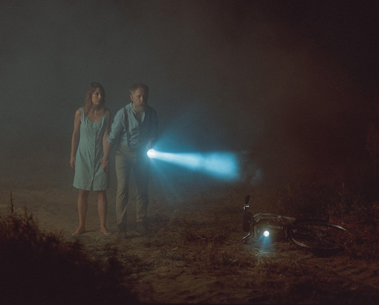 A couple stands on a dirt path at night, holding hands, with the man holding a flashlight and a bicycle lying on the ground with its light on.