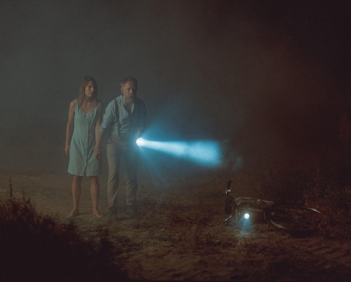 A couple stands on a dirt path at night, holding hands, with the man holding a flashlight and a bicycle lying on the ground with its light on.