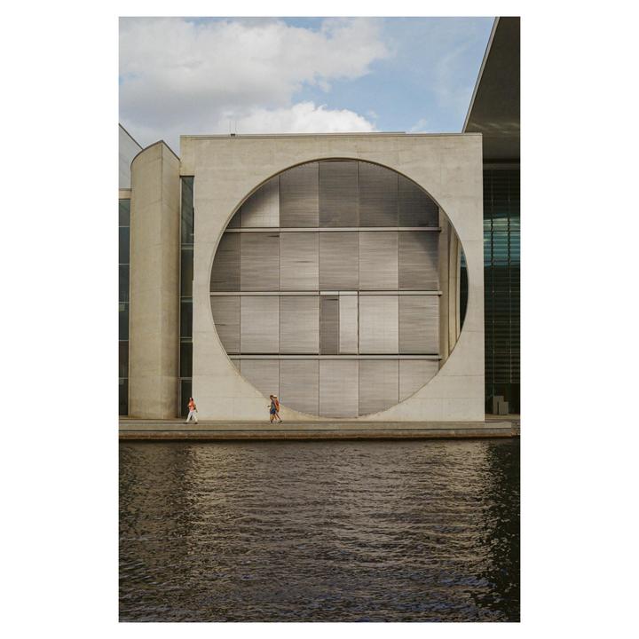 Three people walk along a concrete path next to a body of water, with a large building featuring a circular cut-out in the background.