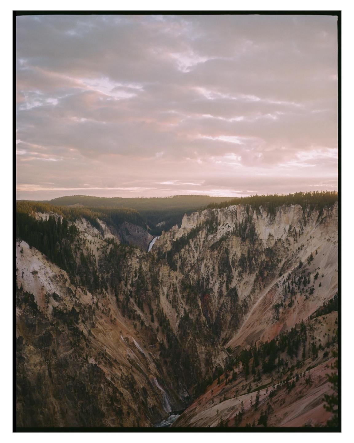 A wide canyon with a river and waterfall is visible under a cloudy sky at dusk.