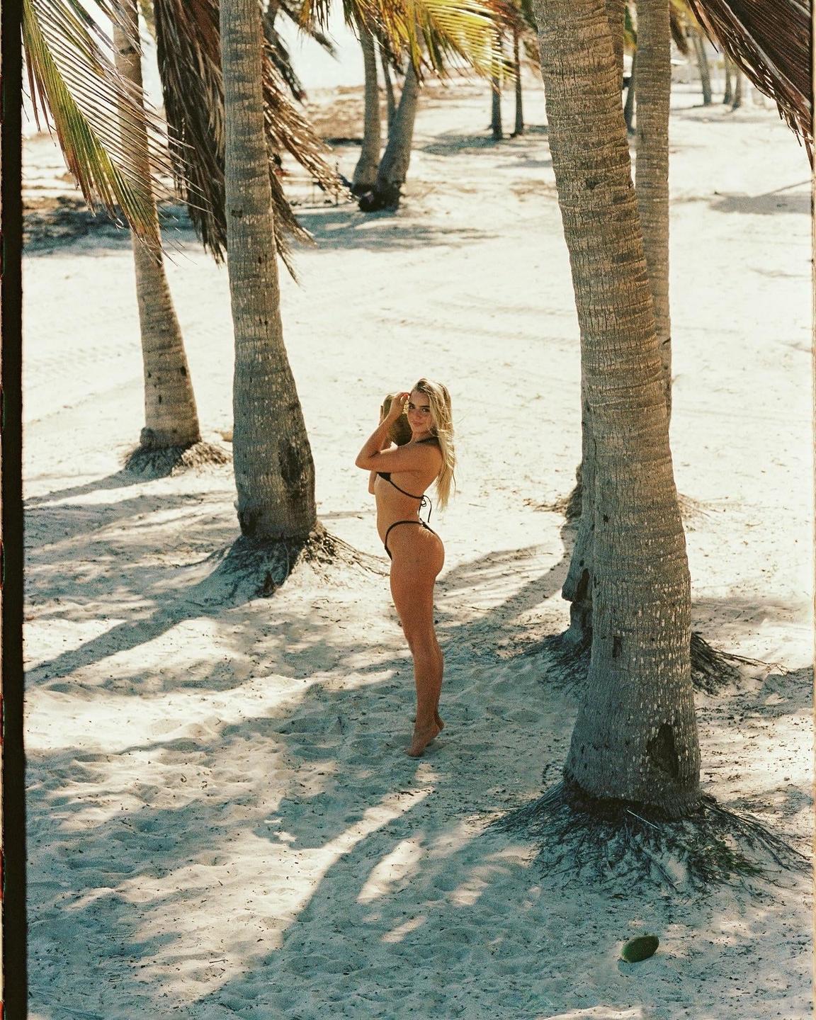 A woman in a bikini stands on a sandy beach among palm trees, holding a coconut.