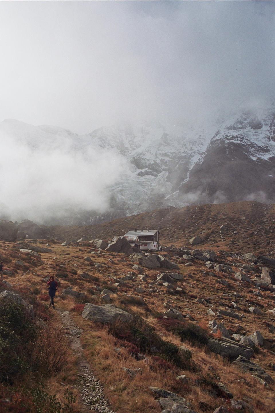 A person walks on a rocky path towards a mountain hut with snow-capped mountains in the background.