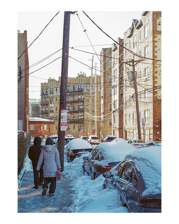 Two people walk on a snowy sidewalk past cars covered in snow and apartment buildings.