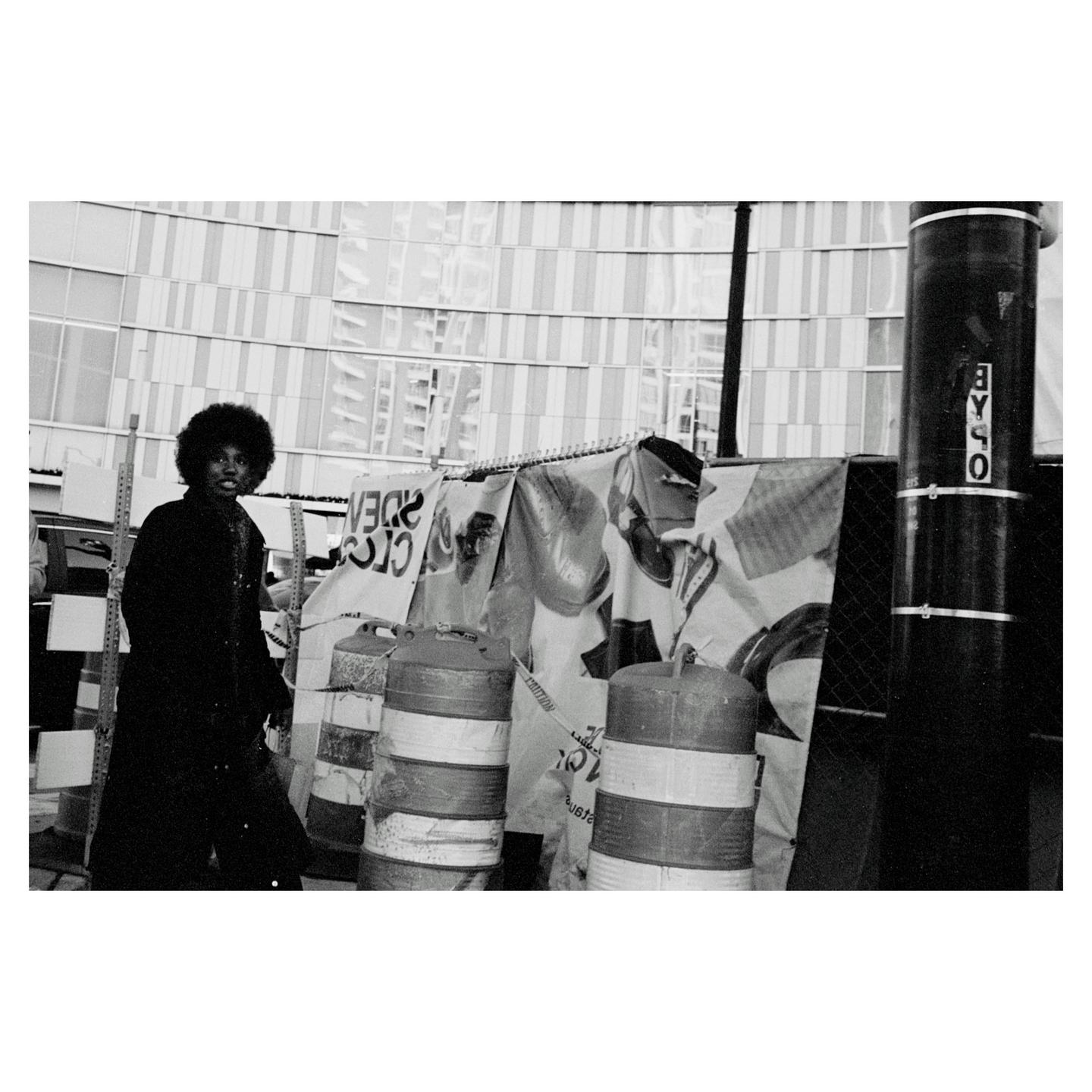 A man with an afro stands on a street next to construction barriers and a building.