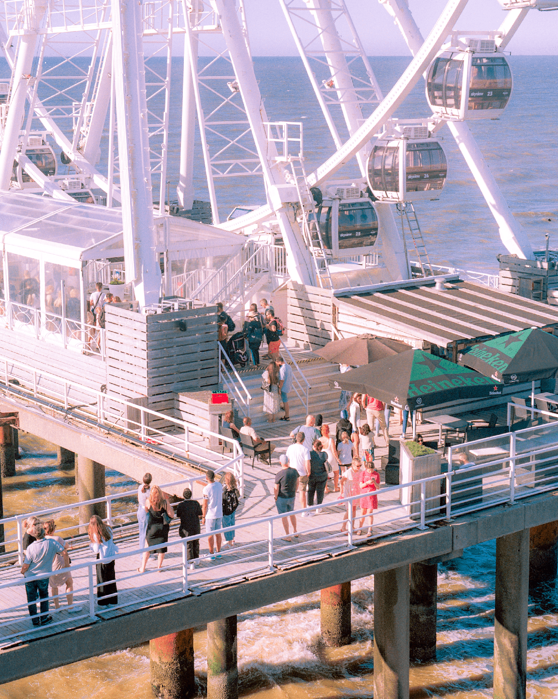 A large white ferris wheel stands over a pier with people walking and standing on it, with the ocean in the background.