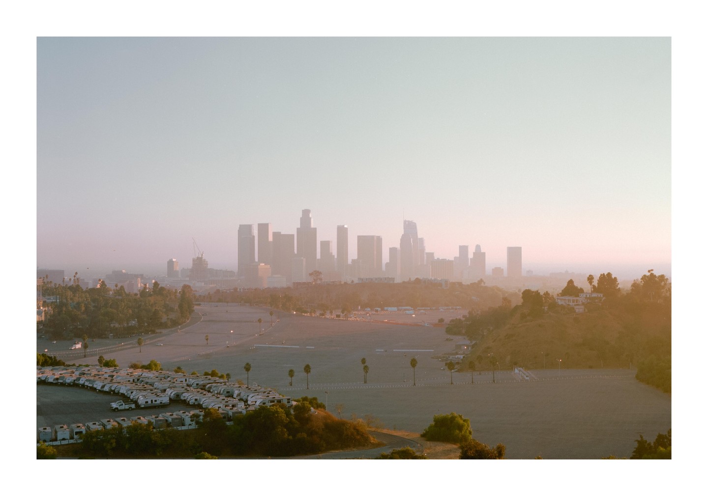 A hazy view of the Los Angeles skyline at dusk, with a large parking lot filled with recreational vehicles in the foreground.