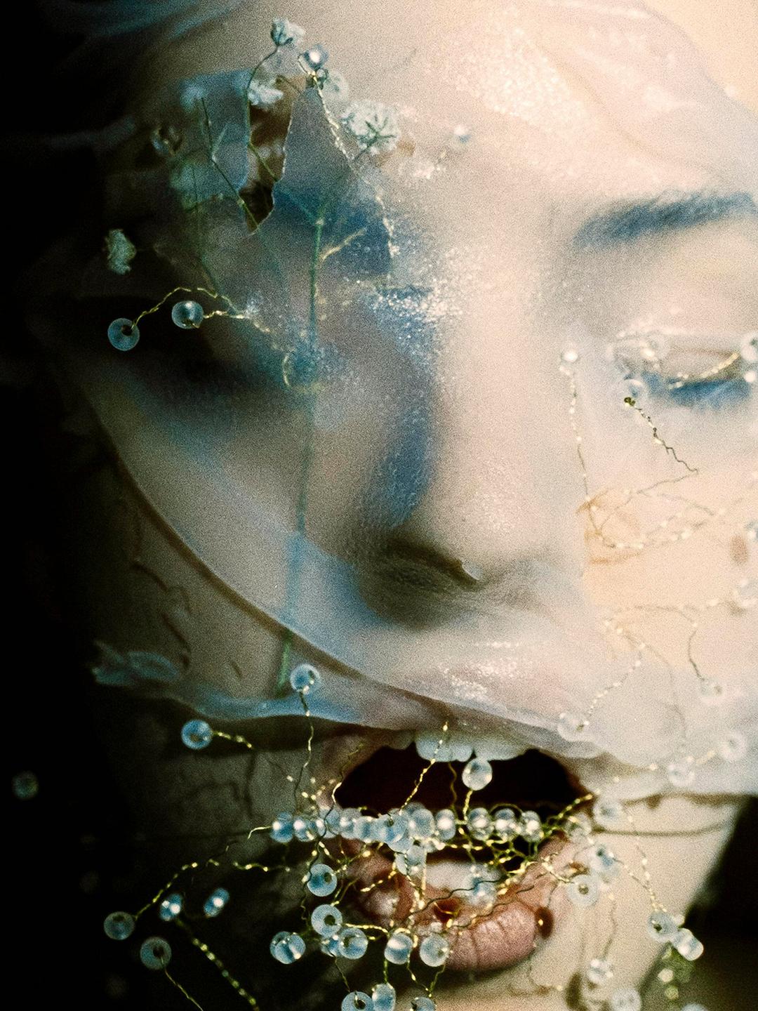 A woman's face is partially covered by a sheer, light-colored fabric adorned with small beads and delicate wire tendrils.