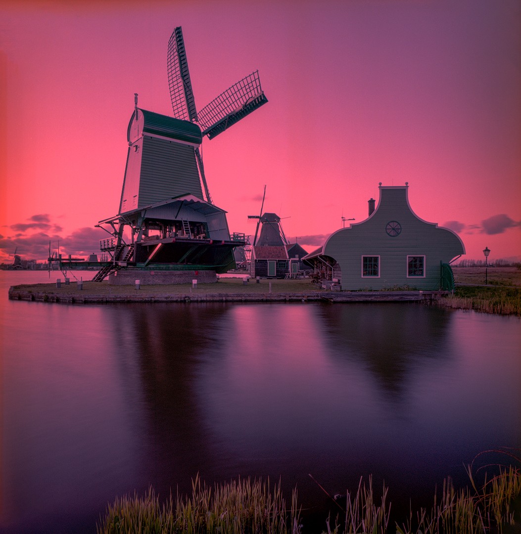 A large green windmill stands next to a body of water with a pink sky in the background.