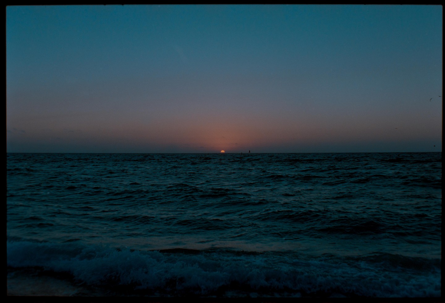 The sun sets over a dark ocean with small waves in the foreground.