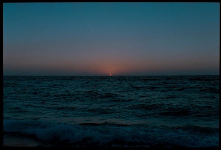 The sun sets over a dark ocean with small waves in the foreground.