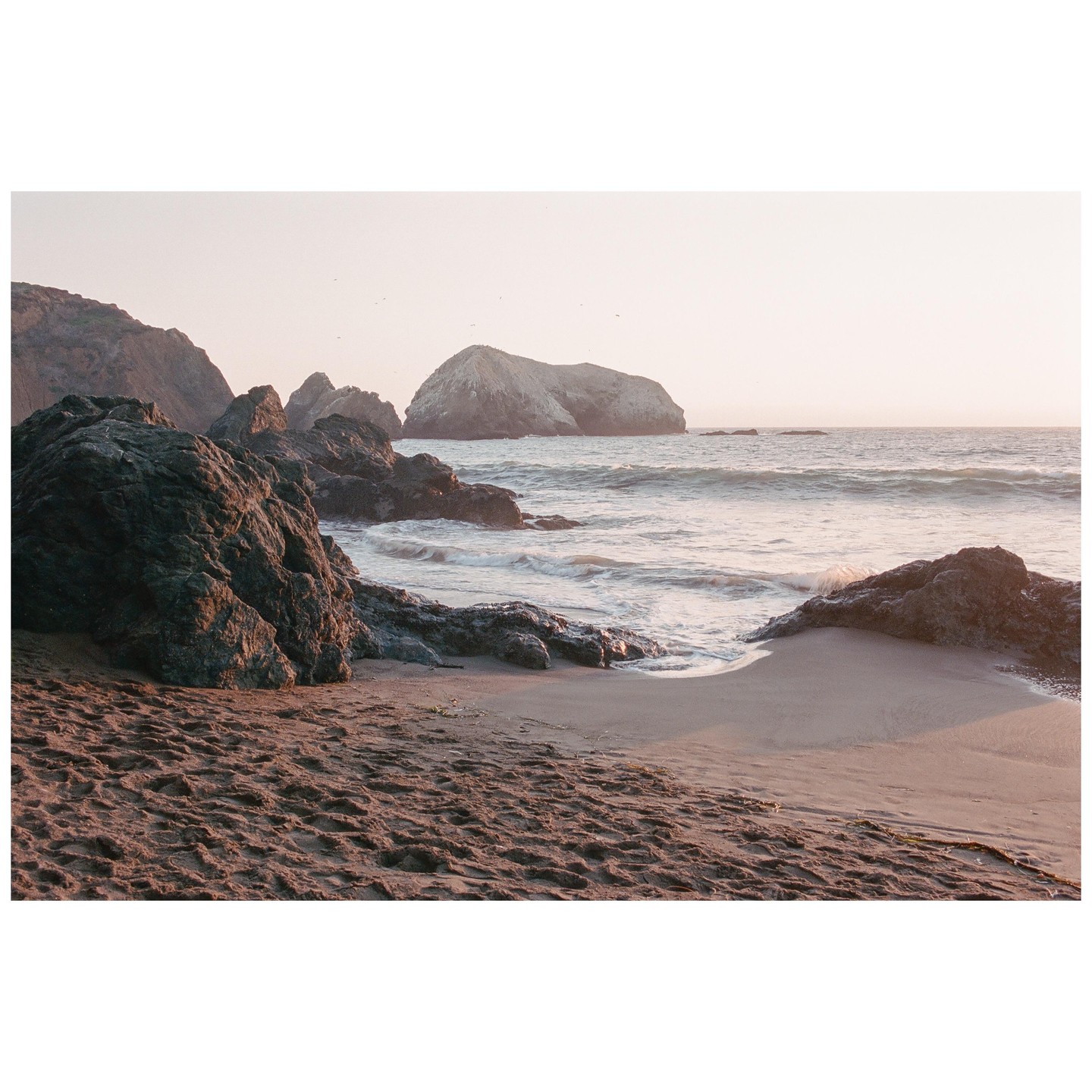 Waves crash against a rocky beach with large rock formations in the distance under a hazy sky.