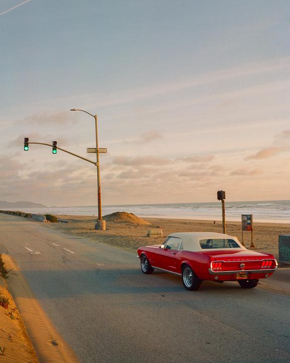A red convertible car is parked on the side of a road next to a sandy beach at sunset.