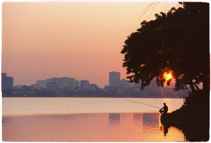 A man fishes from the shore of a lake with a city skyline in the distance during golden hour.