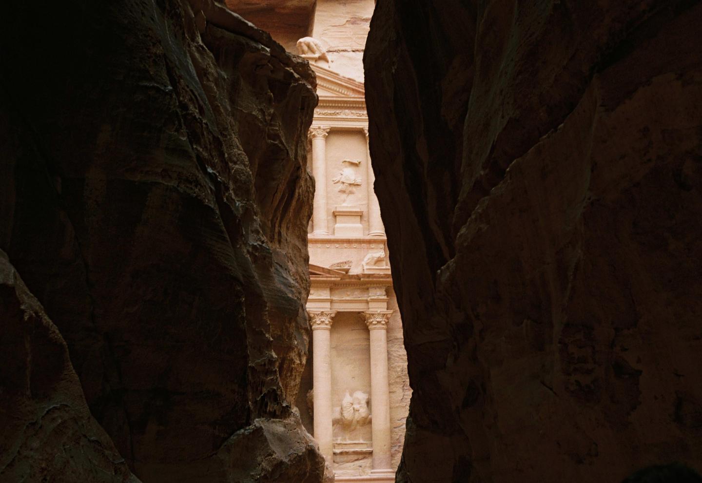A narrow view of a carved stone building is framed by dark rock walls.