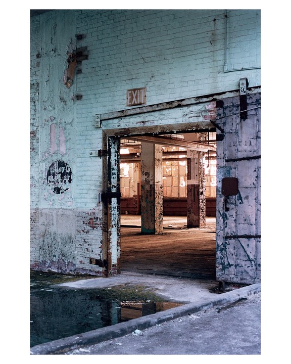 An abandoned factory interior is visible through an open doorway with a puddle of water in the foreground.