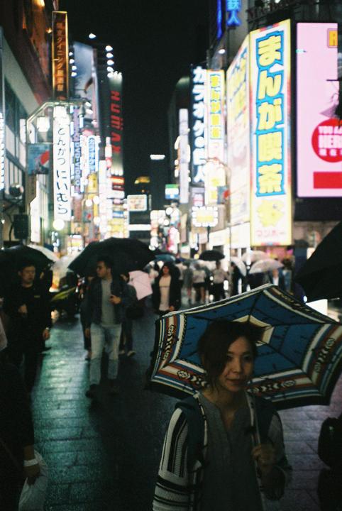 A woman with a blue and white umbrella walks on a wet street at night, surrounded by people and bright neon signs.