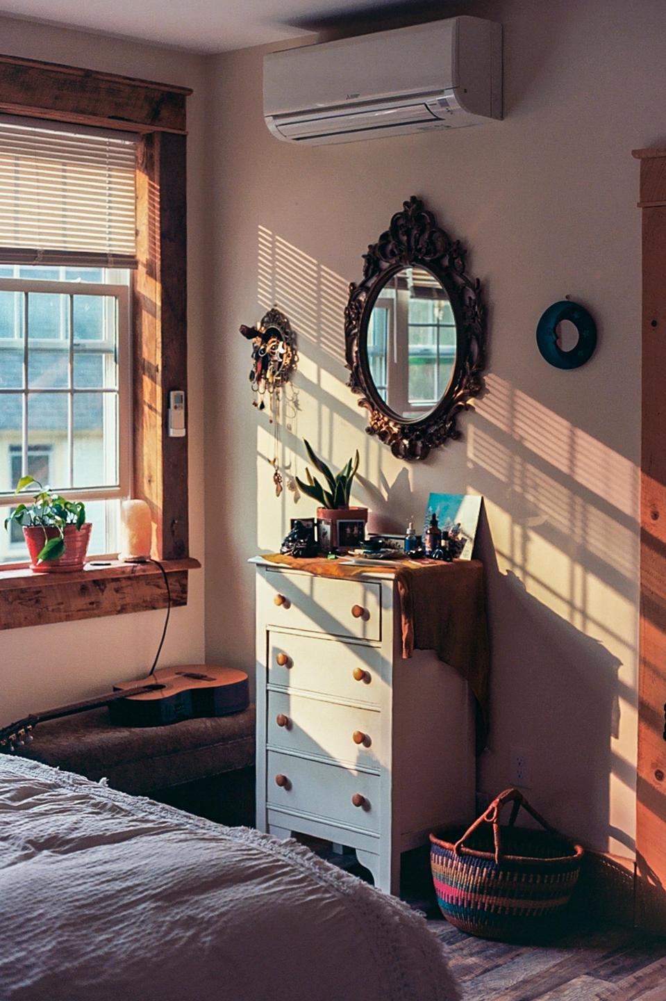Sunlight streams through a window, casting shadows across a bedroom with a dresser, mirror, and guitar.