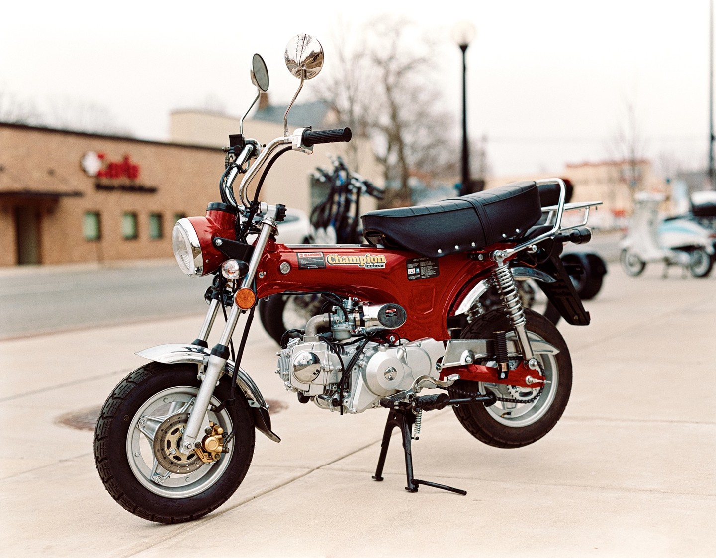 A red Champion motorcycle is parked on a sidewalk with other motorcycles in the background.