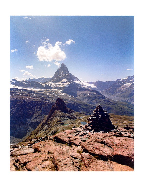 A cairn sits on a rocky mountain peak overlooking a valley with a prominent snow-capped mountain in the distance.