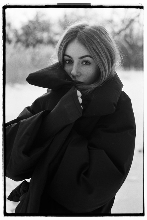 A woman in a large coat looks directly at the viewer.