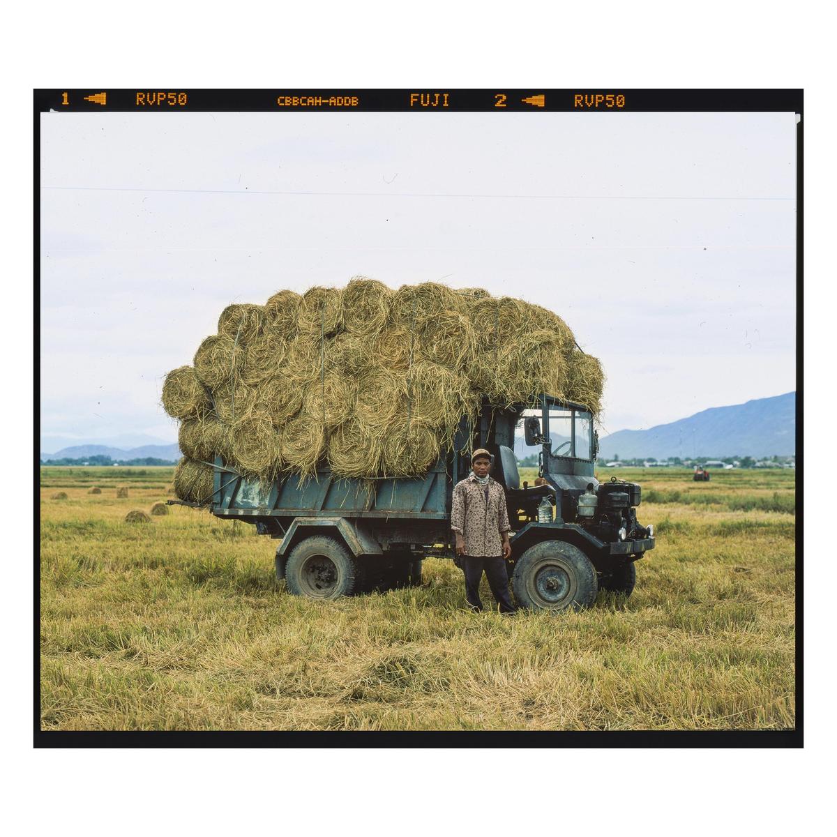 A man stands next to a truck overflowing with hay bales in a field with mountains in the background.