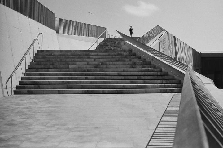 A woman walks up a wide concrete staircase with metal railings and a modern building in the background.