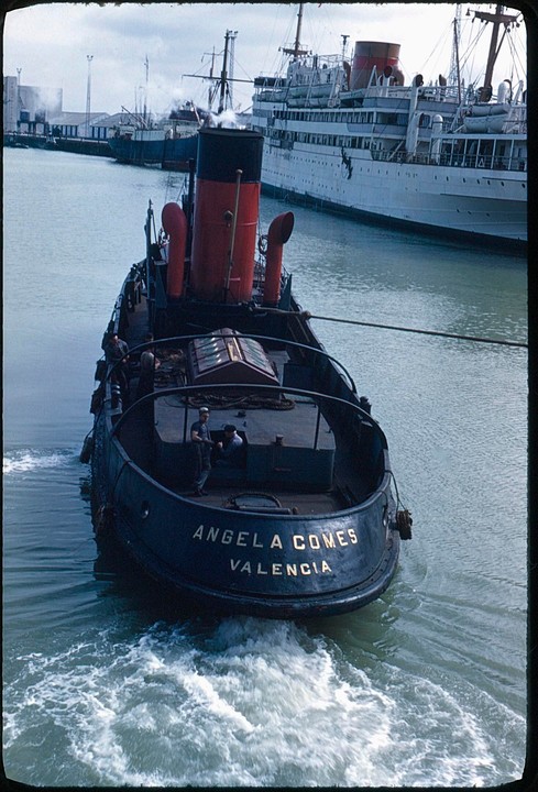 A tugboat named Angela Comes Valencia is pulling a large white ship in a harbor.