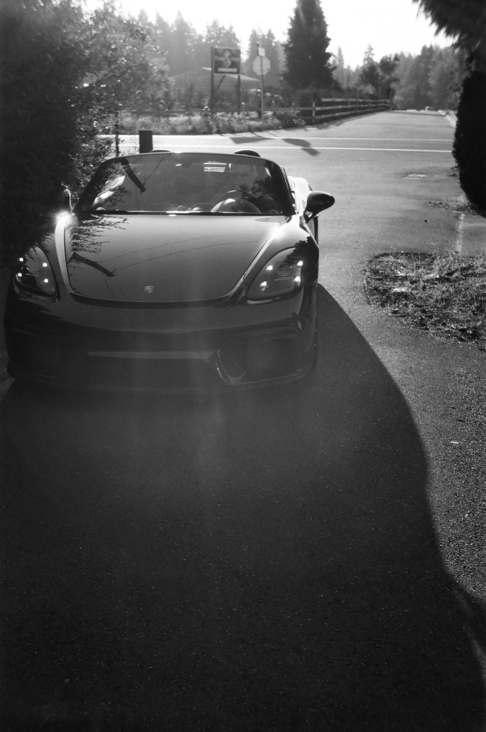 A convertible car is parked on an asphalt road with trees and a fence in the background.