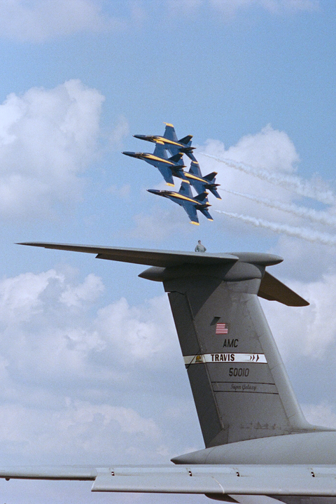 Four blue and yellow fighter jets fly in formation above the tail of a large gray cargo plane.